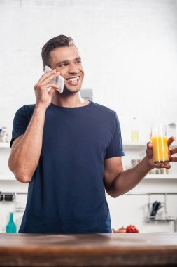 Mutfak masasının yanında portakal suyu bardağı tutarken akıllı telefondan konuşan gülümseyen adam bulanık ön planda. 