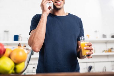 Elinde bir bardak portakal suyu olan gülümseyen bir adamın, mutfaktaki bulanık önplandaki meyvelerin yanında akıllı telefondan konuşması. 