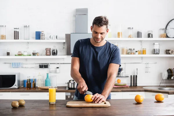 Ung Man Skär Ekologisk Apelsin Nära Kiwi Och Apelsinjuice Köksbordet — Stockfoto