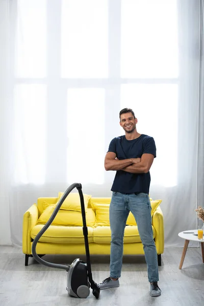 Cheerful man with crossed arms standing near vacuum cleaner at home