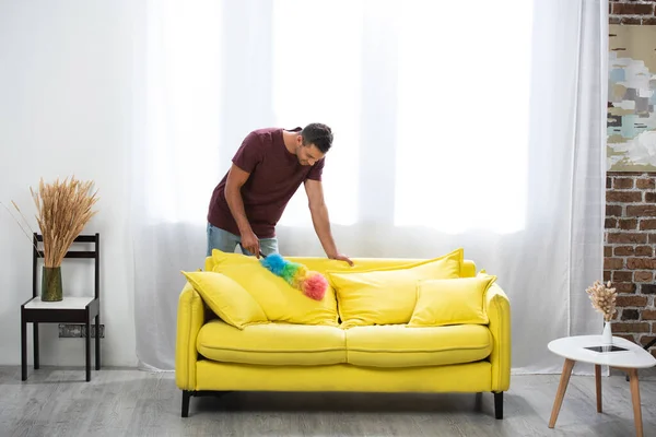 Young man cleaning couch with dust brush near digital tablet on coffee table at home