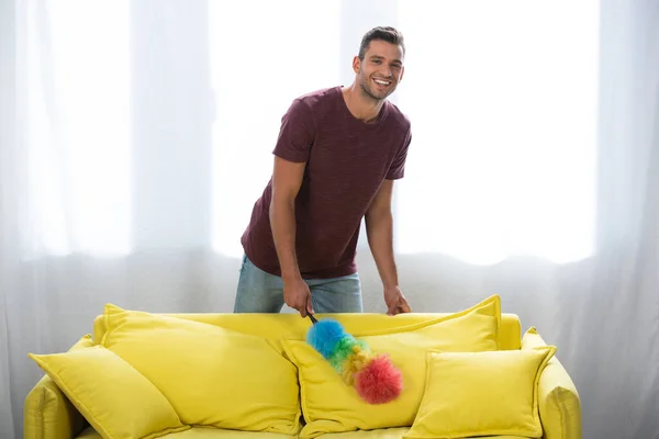 Sorrindo Homem Com Escova Poeira Perto Sofá Amarelo Casa — Fotografia de Stock