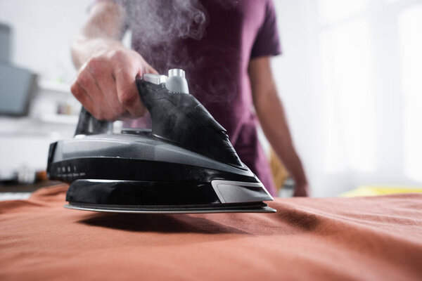 Обрезанный вид железа с паром рядом с одеждой в руке человека на размытом фоне  