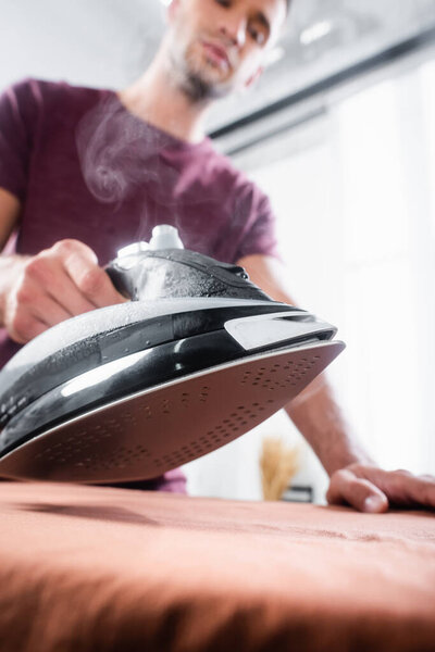 Iron with steam near clothes in hand of man on blurred background 