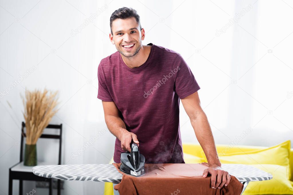 Cheerful man using iron while ironing clothes on board in living room 