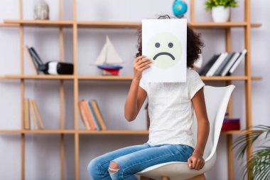 African american girl with autism covering face with unhappy expression on paper while sitting on chair with blurred office on background clipart
