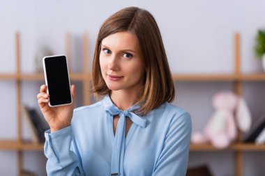 Gülümseyen psikolog kameraya bakarken arka planda bulanık ekranlı akıllı telefonu gösteriyor.