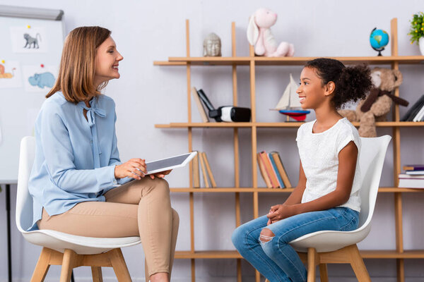 Smiling african american girl looking at psychologist with digital tablet during consultation with blurred office on background