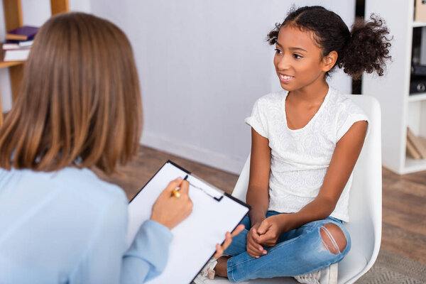 Smiling african american girl looking away while sitting on chair with blurred psychologist with clipboard on foreground