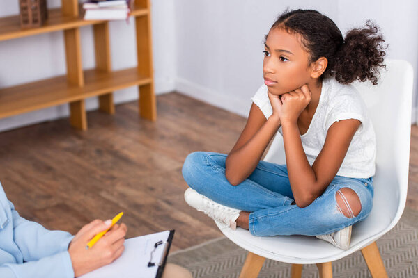 African american girl with crossed legs sitting on chair near psychologist with pen and clipboard on blurred background