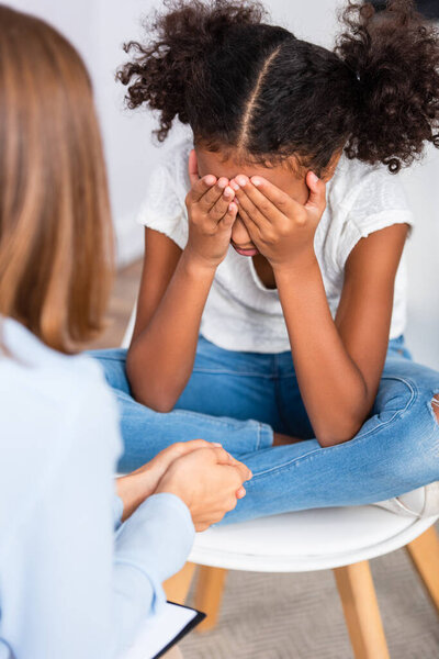 African american girl closing eyes with hands while sitting on chair during consultation with blurred psychologist on foreground