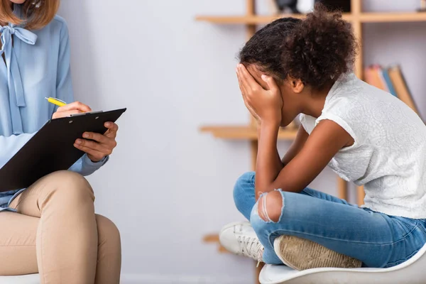 Psicólogo Escribiendo Portapapeles Mientras Está Sentado Cerca Chica Afroamericana Cerrando — Foto de Stock