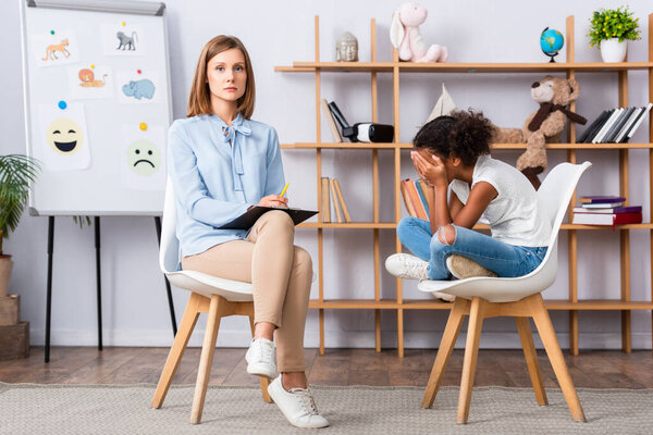 Psychologist looking at camera while sitting on chair near african american girl closing eyes with hands in office