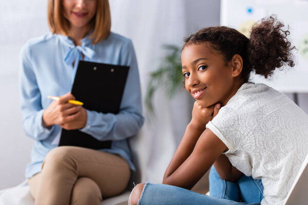 Happy african american girl looking at camera during consultation with blurred psychologist on background