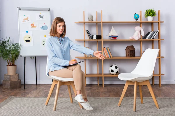 Positive Female Psychologist Clipboard Pointing Hand Empty Chair While Looking — Stock Photo, Image