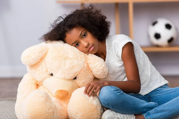 Upset african american girl looking at camera while leaning on teddy bear on blurred background