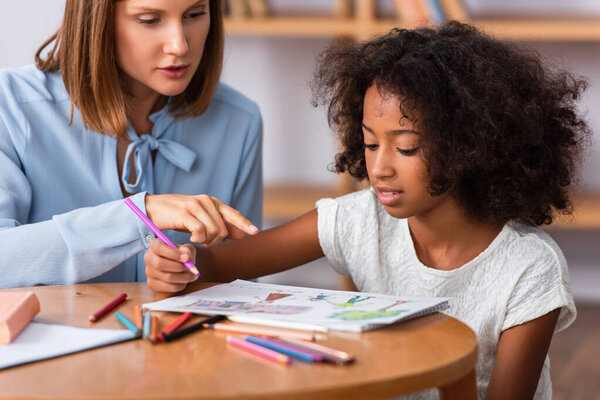 Psychologist pointing with finger at picture near african american girl with colored pencil at coffee table on blurred background