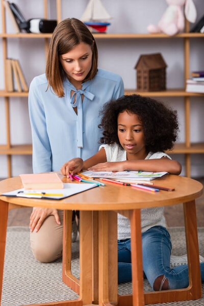 African american girl taking colored pencils on coffee table near psychologist sitting behind on blurred background