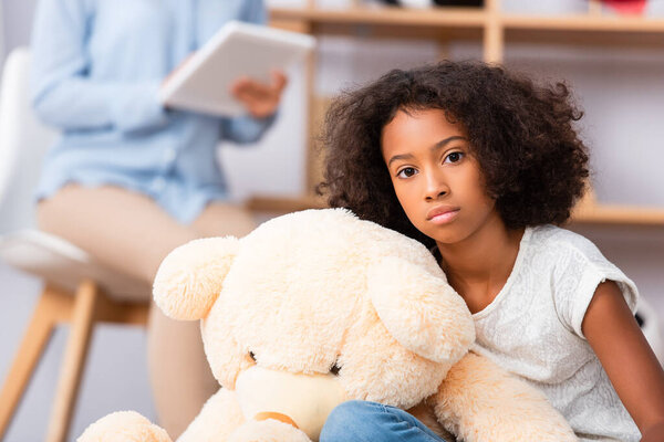 Lonely african american girl looking at camera near teddy bear with blurred psychologist on background