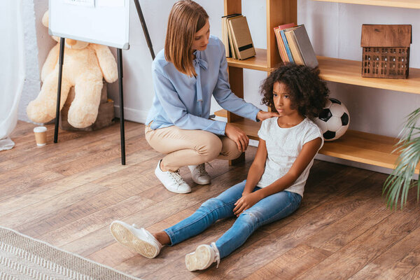 Psychologist touching shoulder of upset african american girl sitting on floor in office