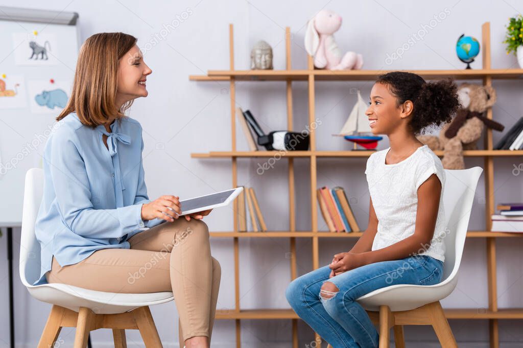 Smiling african american girl looking at psychologist with digital tablet during consultation with blurred office on background