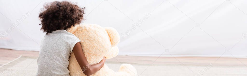 Back view of lonely african american girl hugging teddy bear on floor, banner