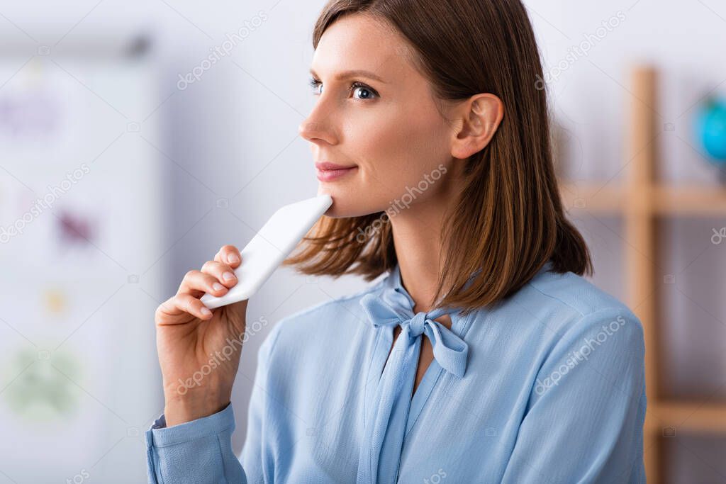 Smiling psychologist with smartphone looking away while thinking on blurred background