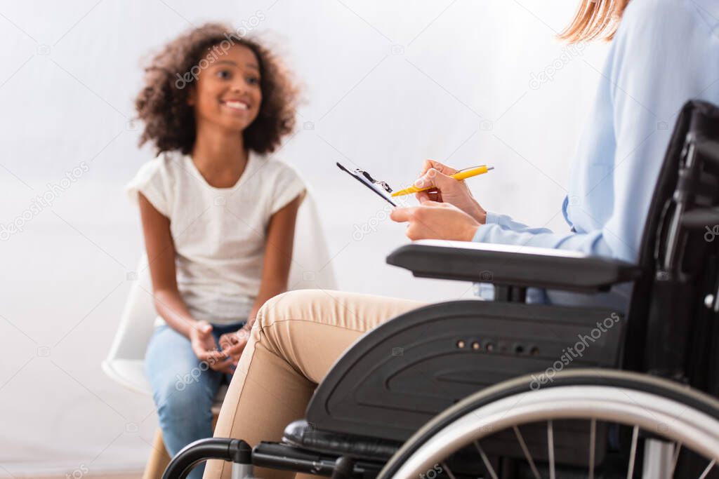 Psychologist writing on clipboard while sitting in wheelchair with blurred happy african american girl on background