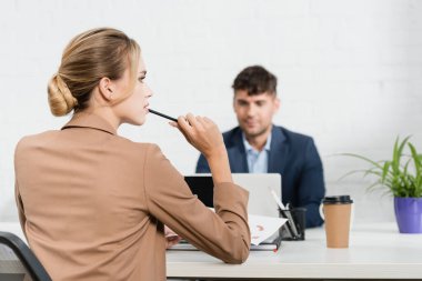 Kalemli düşünceli bir iş kadınının arka planda iş arkadaşıyla birlikte iş yerinde oturması.