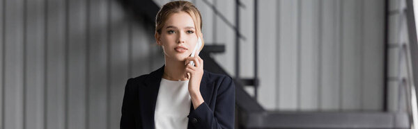 Blonde businesswoman looking away, while talking on mobile phone in office on blurred background, banner