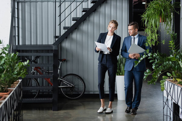 Businessman with laptop looking at paper sheets in hands of female colleague, while standing at work 