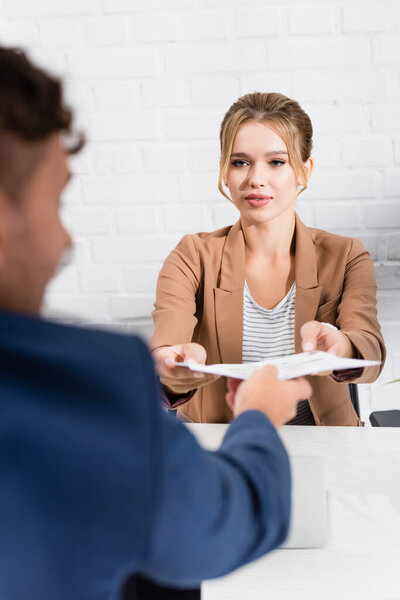 Blonde businesswoman taking document from colleague, while sitting at table on blurred foreground