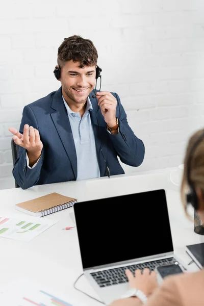 Lächelnder Bediener Gestikuliert Headset Während Arbeitsplatz Mit Verschwommenem Kollegen Vordergrund — Stockfoto