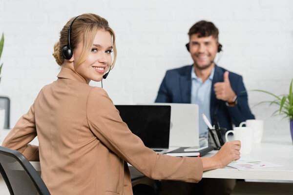 smiling woman in headset looking at camera, while sitting at workplace with blurred colleague on background