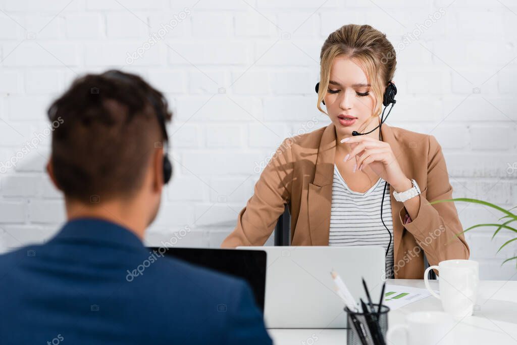 Female operator in headset talking, while sitting at workplace with laptops in office on blurred foreground