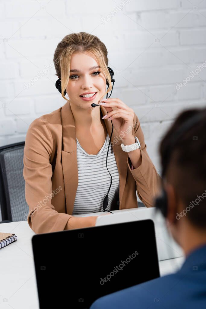 Positive female operator in headset looking at colleague, while sitting at table with digital devices on blurred foreground