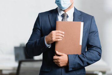 Tıbbi maske takan iş adamının elinde dosyayla görüntüsü bulanık arka planda dururken...