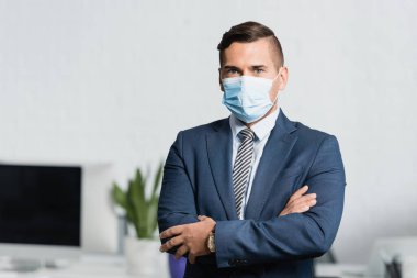 Businessman in medical mask with crossed arms, looking at camera with blurred office on background clipart