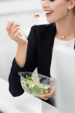 Plastik çatalla yemek yiyen iş kadınının görüntüsü bulanık önplanda yemek kasesini tutarken