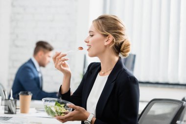 Sarışın iş kadını plastik kaseden yemek yerken iş yerinde bulanık arka planda oturuyor.