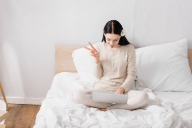 Vitiligo 'su olan ve saçlarında çiçek olan genç bir kadın yatak odasındaki dizüstü bilgisayarında barış işareti gösterirken... 