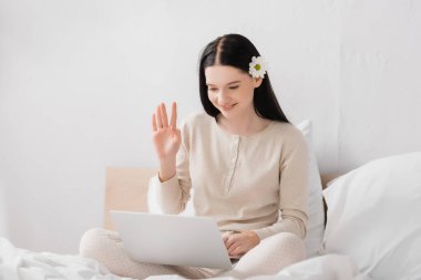 happy woman with vitiligo and flower in hair waving hand while having video call on laptop in bedroom  clipart