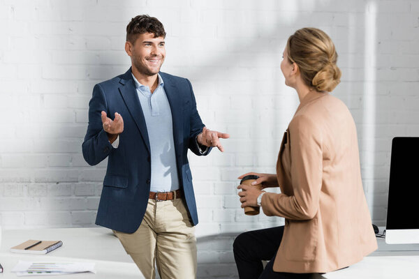 Smiling businessman gesturing, while talking to colleague near workplace during break in office