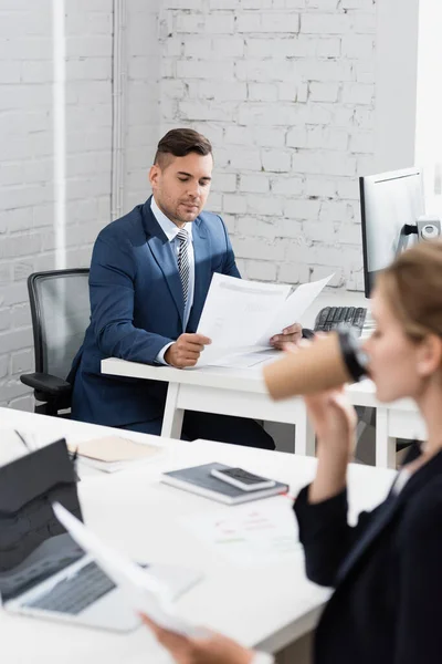 Geschäftsmann Blickt Auf Papierbögen Während Arbeitsplatz Sitzt Mit Verschwommener Kollegin — Stockfoto