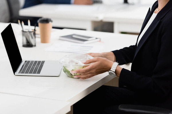 Cropped view of businesswoman holding plastic bowl with meal, while sitting at workplace on blurred background