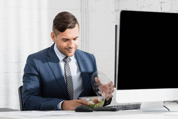 Smiling businessman opening plastic bowl with meal, while sitting at table with computer