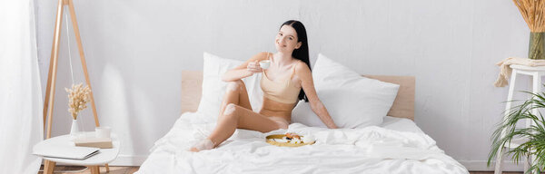smiling woman with vitiligo holding cup near breakfast tray on bed, banner