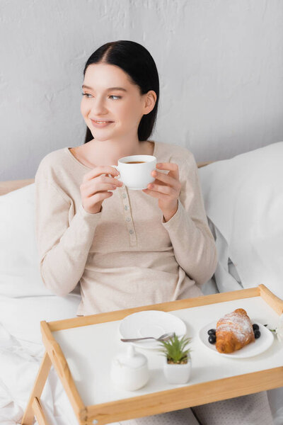 young happy woman with vitiligo holding cup of tea near breakfast on tray