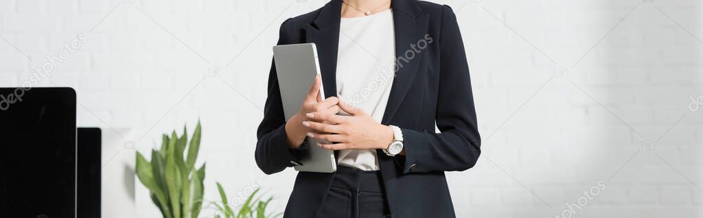 Cropped view of businesswoman in formal wear holding laptop near plants and computer monitors, banner