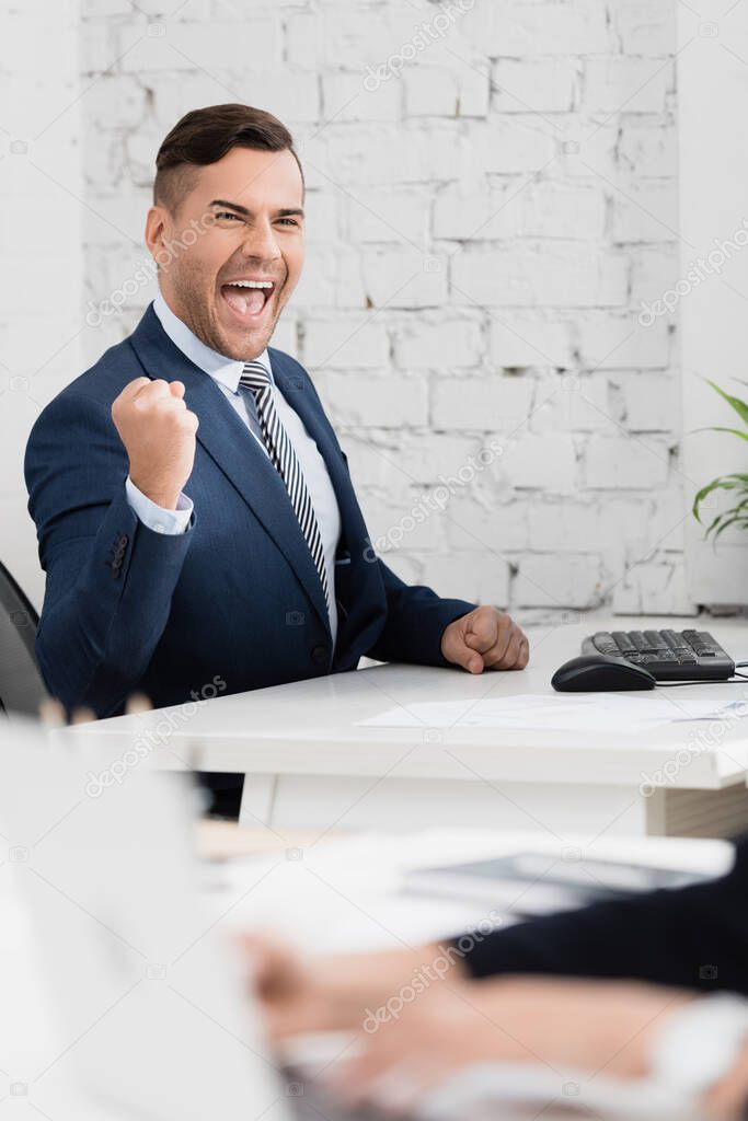 Excited businessman with yes gesture looking at camera, while sitting at table in office on blurred foreground
